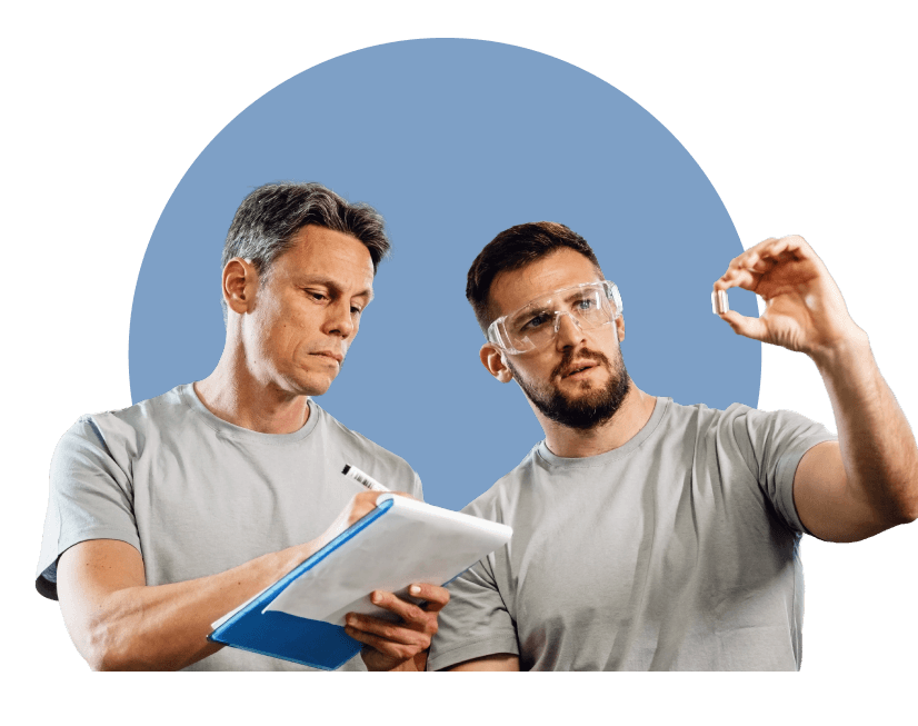 Zwei Personen in grauen Shirts prüfen gemeinsam ein kleines Bauteil; eine Person hält das Bauteil mit Schutzbrille in der Hand, die andere macht Notizen auf einem Klemmbrett.“